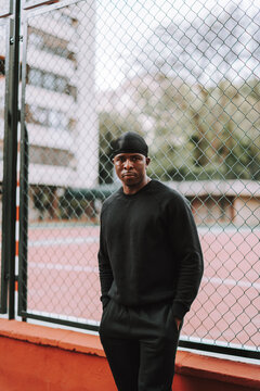 Chico negro apuesto vestido totalmente de negro posando delante de una cancha de baloncesto con reja