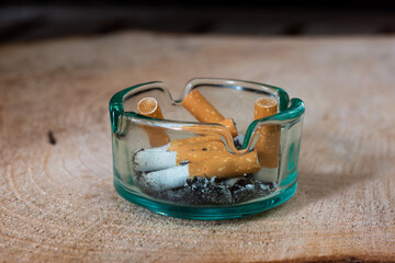 Clear glass cigarette ashtray with butts and ash set on wooden board close up macro shot shallow depth of fie