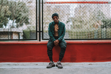 Chico negro joven y guapo posando junto a una cancha de baloncesto entre edificios