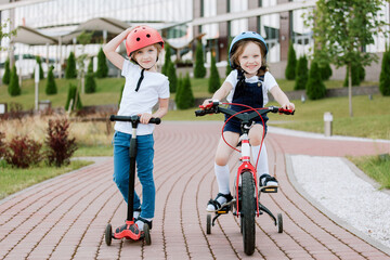 Two little girls having fun on bicycle and scooter. Cheerful sisters in helmets riding outdoors.
