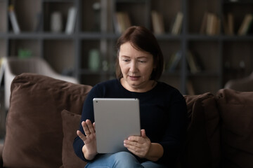 Modern elderly Caucasian 60s woman sit relax on sofa at home use tablet gadget talk speak on video call online. Smart mature grandmother have webcam digital virtual conference on pad.