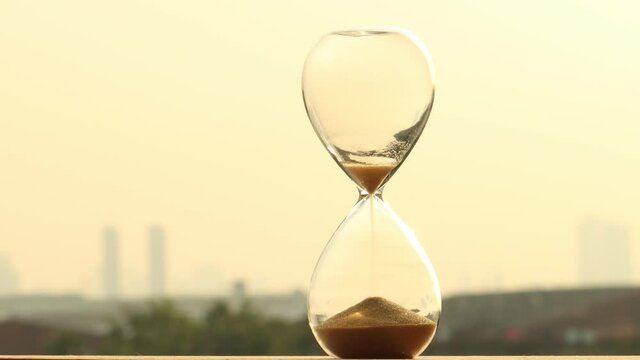 Sand clock measuring time with orange sky in evening time over city blurred background, yellow sand falling down in hourglass old classic timer.