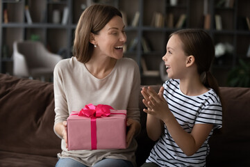 Cute little teen girl child present beautiful handmade wrapped gift to excited young Caucasian mother for Mothers Day. Happy small teenage daughter congratulate greet mom with birthday make surprise.