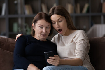 Stunned adult kid child and elderly mother relax at home shocked with promotion deal or discount sale on smartphone. Amazed grownup daughter and old 60s mom feel surprised with news on cellphone.