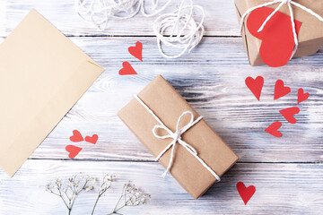 Gift in craft paper packaging with paper red hearts and envelope on a white wooden background,...