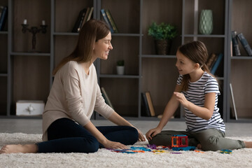 Little teen girl child and young Caucasian mother or nanny sit on floor at home play on weekend together. Happy mom and small teenage daughter have fun engaged in funny game activity in living room.