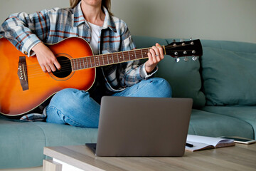 Music college hipster student in checkered shirt practicing acoustic guitar exercise, reading notes from laptop. Young woman taking an online musical courses, practicing at home. Background, close up