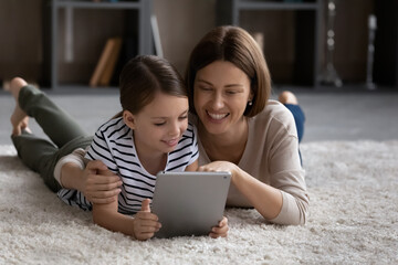 Loving happy young mother and small teenage daughter relax in living room talk on video virtual webcam call on pad. Happy mom and little girl child rest have fun using modern tablet gadget at home.