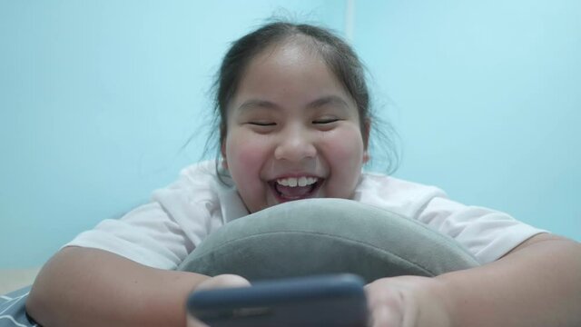 Happy chubby cute Asian girl watching online video, video call, message, cartoon, play game in smartphone. Adorable kid lie prone on pillow in home was smiling and laugh a lot. Front view