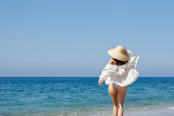 Caucasian woman with fit body wearing loose white cotton shirt blue bikini bathing suit and broad brim straw hat posing at sandy beach on beautiful sunny day. Mediterranean sea background. Copy space