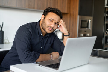 Depressed unemployed hindu man is using laptop for job seeking, upset pensive young indian guy sits in the kitchen at home with the laptop, rested head with hand, looks away, feels sad and worrying