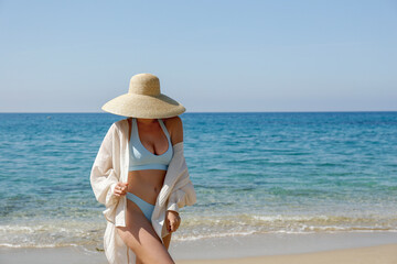Fototapeta na wymiar Caucasian woman with fit body wearing loose white cotton shirt blue bikini bathing suit and broad brim straw hat posing at sandy beach on beautiful sunny day. Mediterranean sea background. Copy space