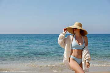 Caucasian woman with fit body wearing loose white cotton shirt blue bikini bathing suit and broad brim straw hat posing at sandy beach on beautiful sunny day. Mediterranean sea background. Copy space
