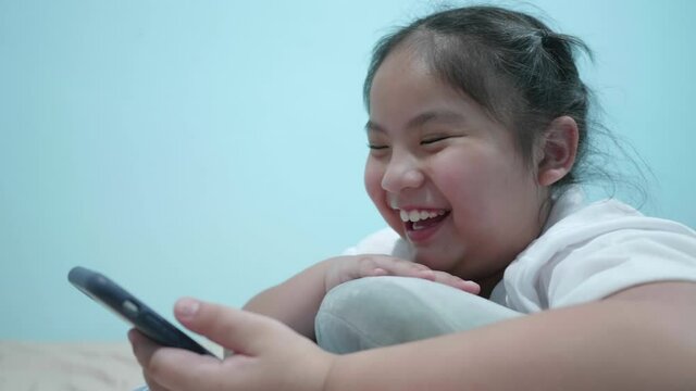 Happy chubby cute Asian girl watching online video, video call, message, funny cartoon in smartphone. Smile adorable kid lie prone on pillow in home was laugh a lot and point fingers to screen