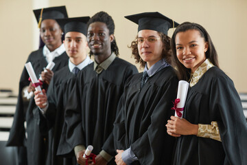 Multi-ethnic group of happy young people wearing graduation robes standing in row and looking at camera