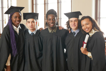 Waist up portrait of multi-ethnic group of young people wearing graduation robes and smiling at camera while standing in school auditorium