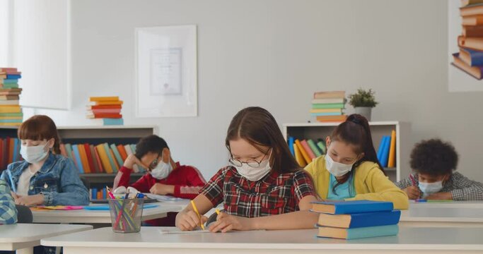Diverse children raising hands for answer wearing medical masks for protection from virus at lesson