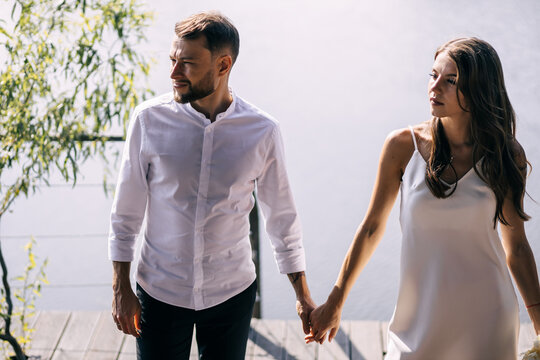 Newlyweds at the pier. The bride and groom walk along the pier to hold hands, in fashionable clothes near a green tree, pensive look to the side.