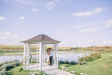 Lovely young couple hugging in a summer white gazebo near the lake with a beautiful landscape.