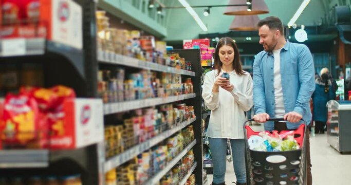 Sweet young family choosing products for cooking dinner together in supermarket. Caucasian couple with shopping cart buying groceries and talking to each other. Marriage, purchase concept.
