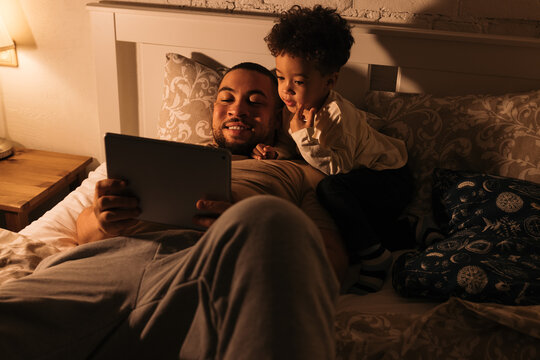 Smiling Dad And His Little Son In Bed Watching Content On A Digital Tablet