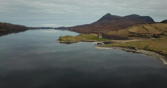4k aerial footage of ruins of Ardvreck Castle and Loch Assynt with mountains in background. Clouds and sky reflected in calm water of loch. Panning backwards away from castle.