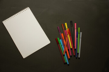 Markers of different colors with a white notebook on a black background. View from above. Colorful pens with a close-up notebook. Education, art, creativity. The markers are on the right. Copy space.