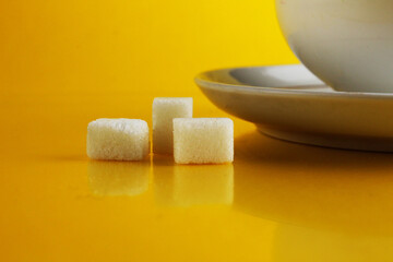 A white cup on a saucer with tea or coffee next to lie cubes of sugar slices and a doughnut in the glaze on a bright yellow background with a place for a copyspace. Still life with sweet food