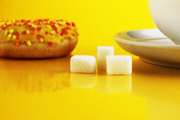 A white cup on a saucer with tea or coffee next to lie cubes of sugar slices and a doughnut in the glaze on a bright yellow background with a place for a copyspace. Still life with sweet food