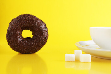 A white cup on a saucer with tea or coffee next to lie cubes of sugar slices and a doughnut in the glaze on a bright yellow background with a place for a copyspace. Still life with sweet food