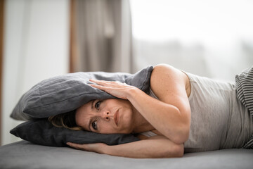 Sleepless lady covering ears with pillow. Noisy neighbors, tinnitus, insomnia or stress concept. Tired woman can't sleep. Awake in bed after coming home from her shift, work.