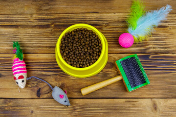 Obraz na płótnie Canvas Dry cat food in bowl, cat toys and pet slicker brush on wooden background. Top view. Pet care concept