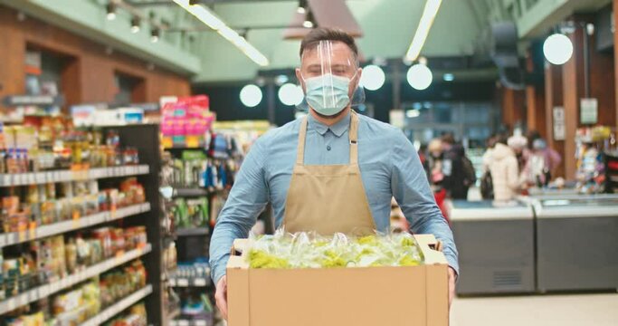 Successful male store owner wearing face visor, carrying box with lettuce in supermarket. Adult Caucasian man in medical mask holding vegetables. Shelves of goods, customers in background. Commerce.