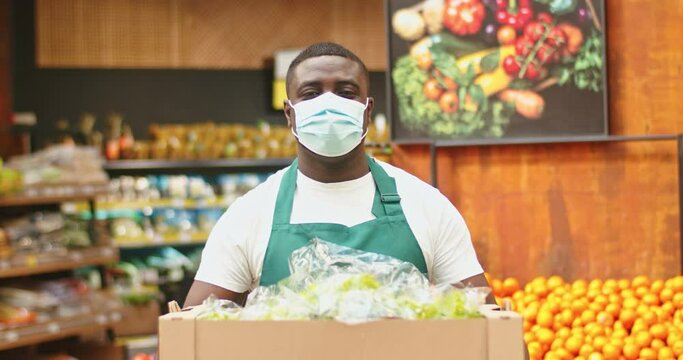 Portrait of attractive male saller in apron holding box full of lettuce in grocery store. Young Afro-American man wearing medical mask looking at camera at vegetables, fruits section. Virus, health.