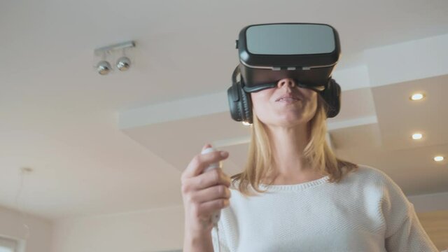 Middle shot of a smiling Caucasian woman using the head-mounted display at home, playing games, holding VR controllers