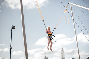 A girl of about 8 years old is jumping on a bungee trampoline. A child with a harness and elastic bands hangs against the sky. The concept of a happy childhood and games in an amusement park by the se
