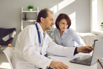Young woman listening to senior doctor and looking at health screening results on laptop computer screen. Mature medical specialist at modern clinic showing X-ray scans to middle-aged patient