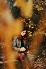 Pensive red-haired woman in the park on wooden bench. Pretty woman in beret and leather jacket. A person with a graphic tablet, a designer, a freelancer. Autumn portrait.