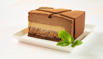 A slice of chocolate modern cake on a square white plate with a piece of green mint with a beautiful cut, on which layers of mousse are visible