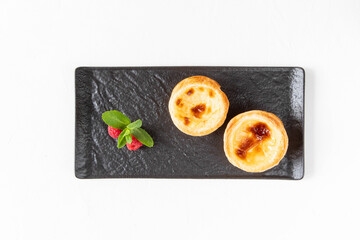 Two Portuguese Pastel de nata cakes, consisting of egg yolk custard on a black plate - dessert for morning breakfast with fresh berries - top view