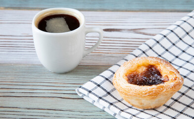 Traditional dessert in Portugal - Pastel de nata cake on textiles on a wooden table with a cup of hot black coffee in a white ceramic - side view