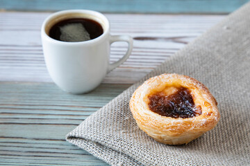 Traditional dessert in Portugal - Pastel de nata cake on textiles on a wooden table with a cup of hot black coffee in a white ceramic - side view