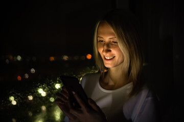 Portrait of a beautiful young blonde woman with an open smile in a white shirt with a mobile phone in her hands near the window of a night city background, warm light from the phone falls on her face