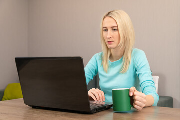 Fototapeta premium Young blonde woman is sitting at a table with a laptop, reading the news, drinking tea or coffee from a green mug
