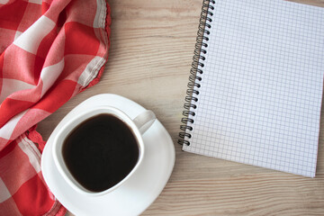 White cup of strong black coffee with red-white cloth and notepad on wooden surface close up