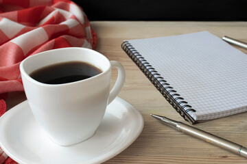 White cup of strong black coffee with red-white cloth, metal pen and notepad on wooden surface close up