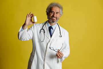 Old doctor shows pack of capsules pills. Portrait of physician advertising pills. Yellow background.