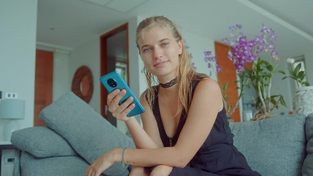 Gorgeous young blonde woman sitting on a sofa wearing sexy black dress with choker on her neck and using mobile phone looking at dating apps as she swipes and sends seductive messages
