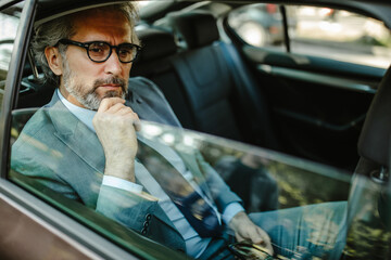 Senior businessman driving on car back seat, looking away while holding a smart phone.