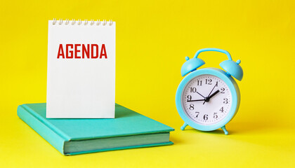 The word AGENDA is written on a notebook that lies on a green diary, next to an alarm clock on a yellow background